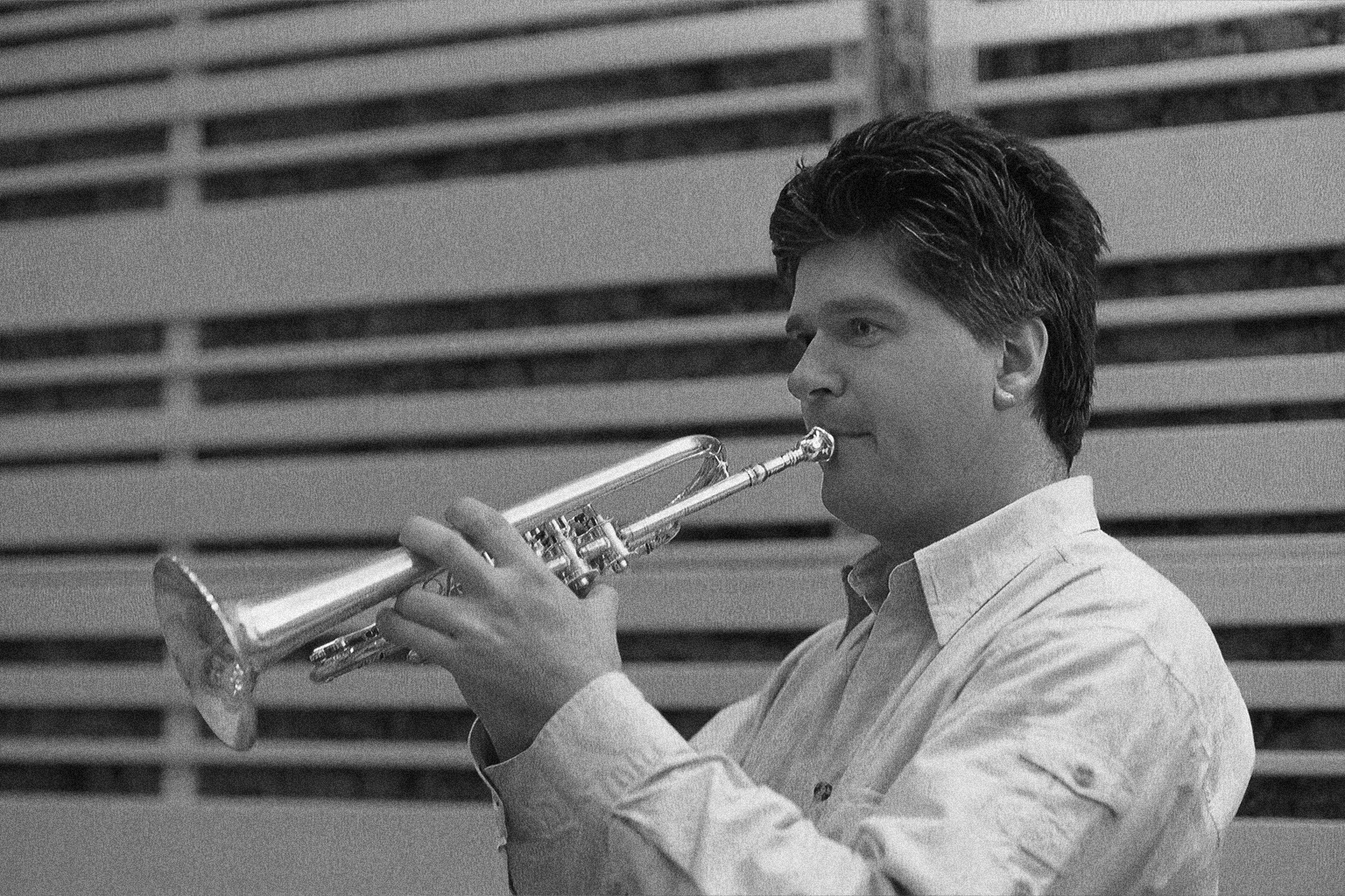 Black and white image of a man playing a shiny trumpet, his fingers positioned on the valves, with a striped background.