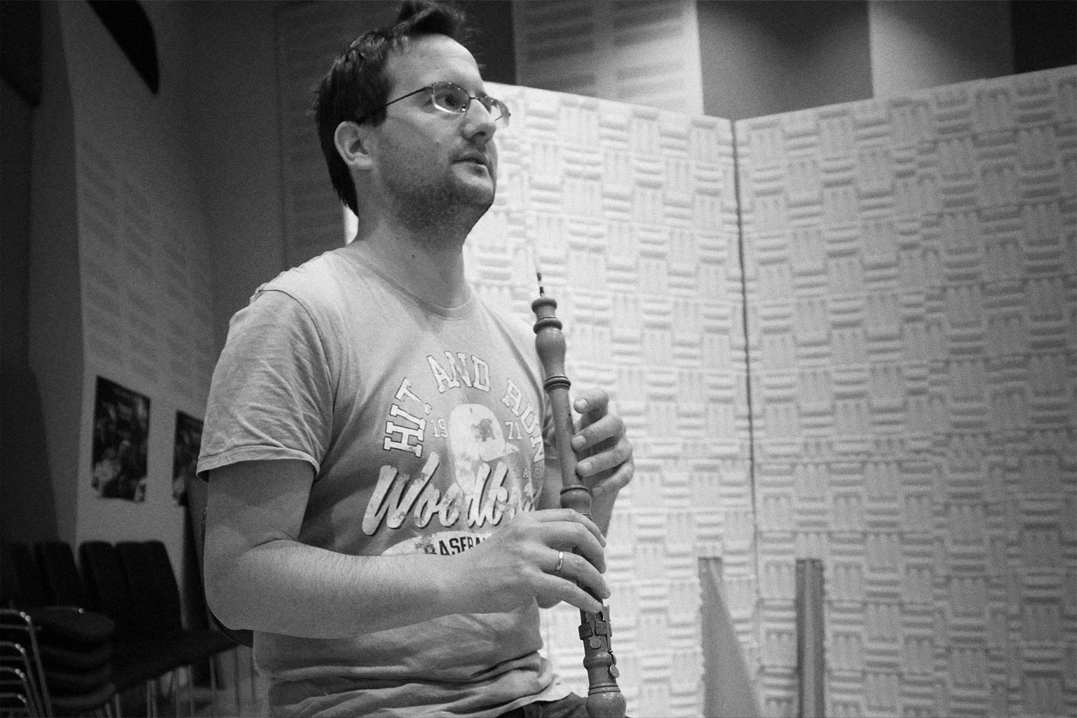 Black and white image of a man in glasses holding a woodwind instrument, possibly an oboe or bassoon, in a music studio with soundproofing panels.