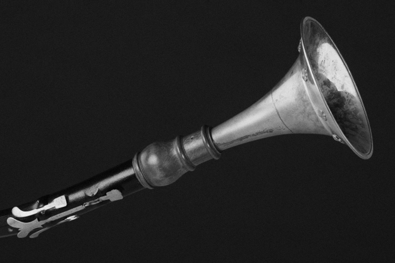 Black and white photo of a vintage wind instrument, possibly a cornet or horn, with a metallic bell and a dark body featuring keys, set against a plain dark background, highlighting its antique musical design