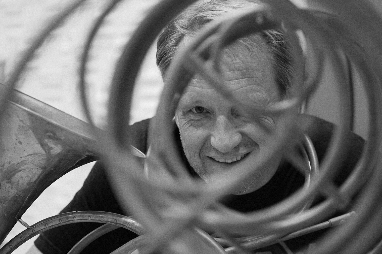 Black and white close-up of a smiling man with a beard looking through the curved metal piping of a tuba, his eye visible through a gap in the tubing, with the rest of the instrument and a black shirt also visible.