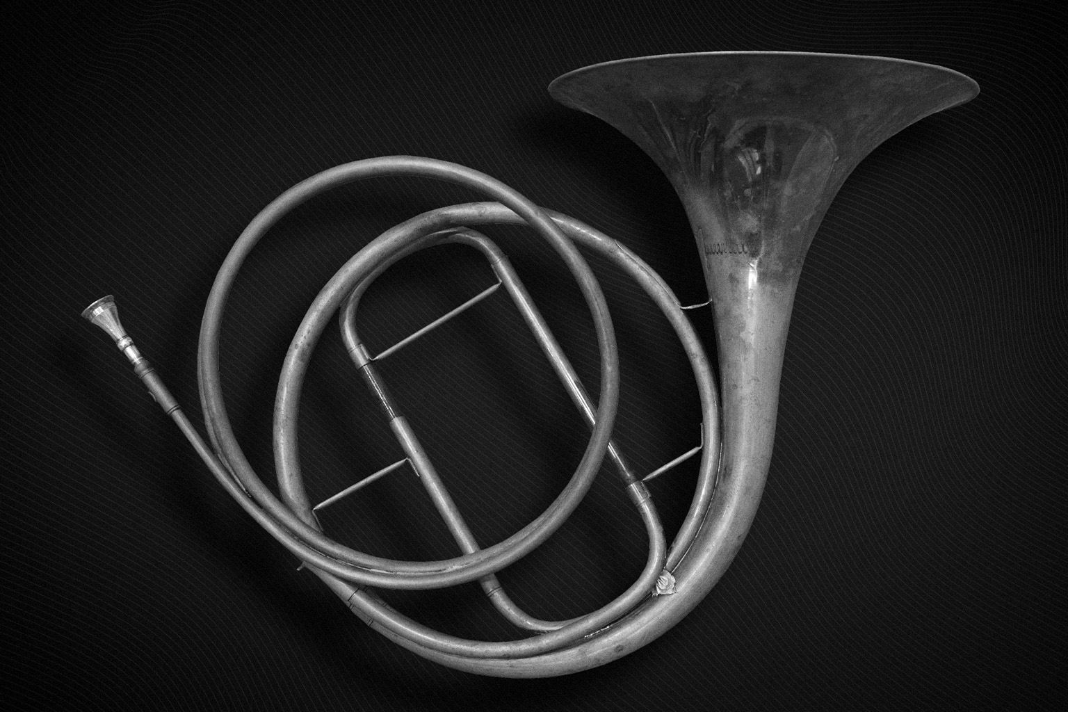 Black and white photo of a French horn with its bell and coiled tubing prominent, set against a dark, textured background.