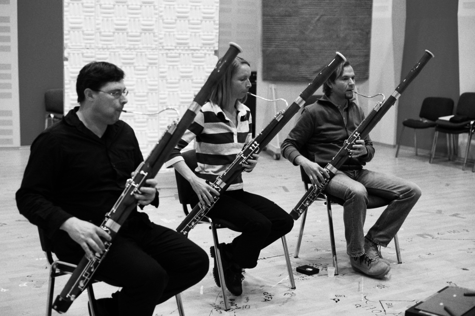 Black and white image of three musicians playing bassoons, seated in a rehearsal space with soundproofing panels.