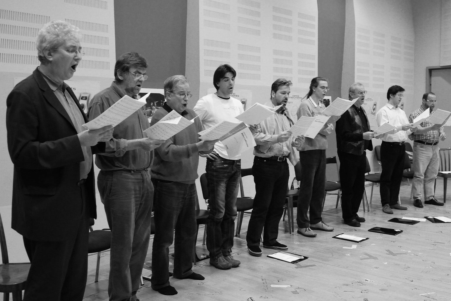 Black and white image of a male choir practicing. Nine men stand in a row, holding sheet music and singing, mouths open. There are chairs behind them and scattered papers on the wooden floor.