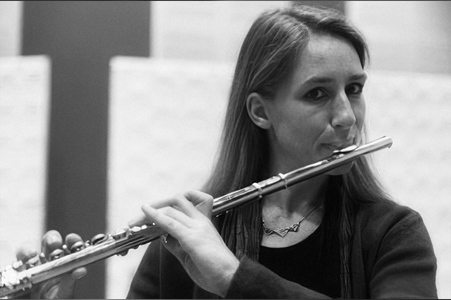 Black and white photo of a long-haired woman playing a flute, her fingers pressing the keys, with a blurred background featuring a dark T-shaped structure.