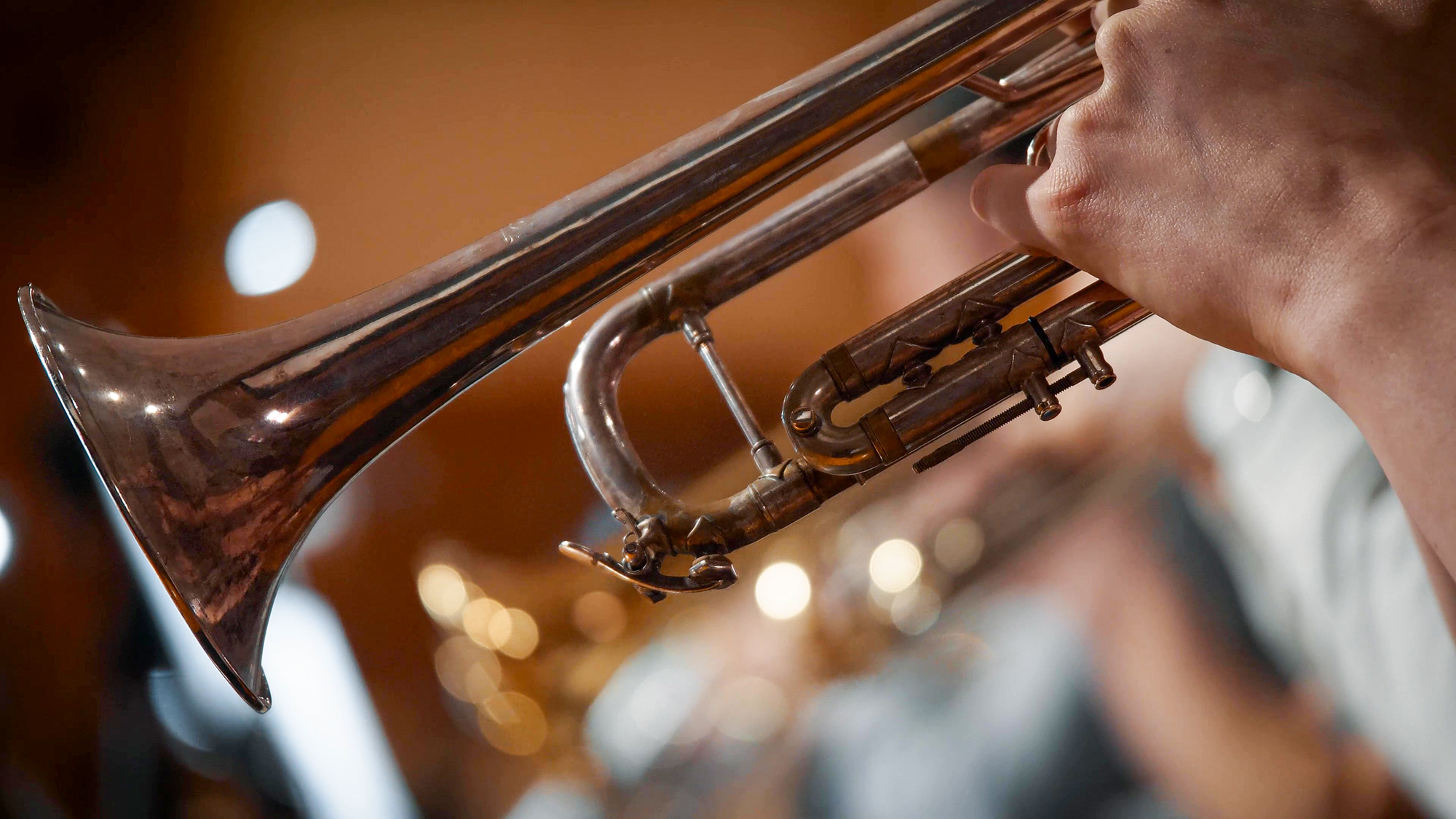 Close-up of a person's hand holding and playing a brass trumpet with a blurred background of lights and musical instruments.