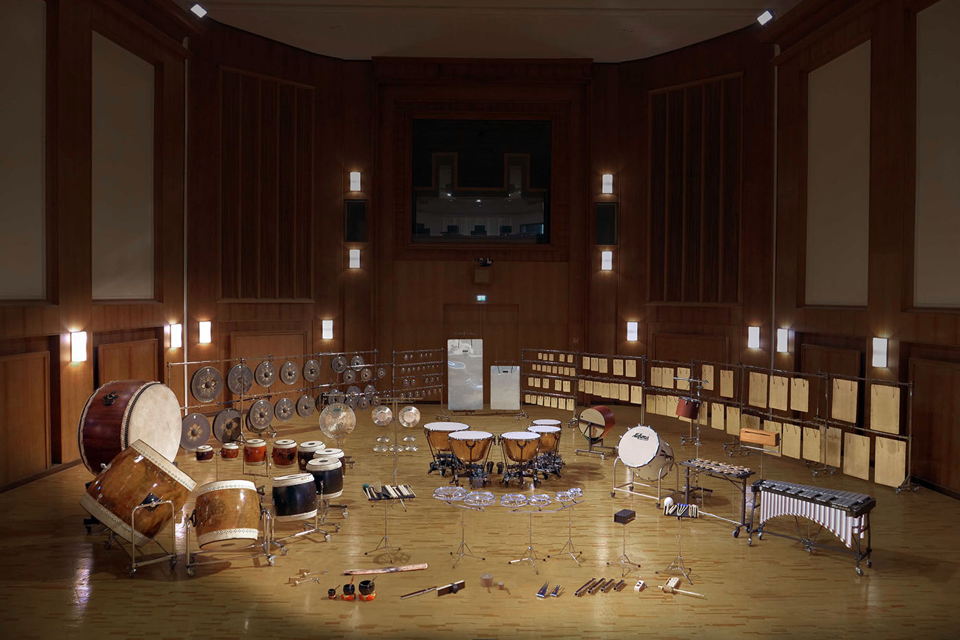 Orchestral percussion instruments including drums, gongs, timpani, cymbals, bass drum, and xylophone arranged on a wooden stage in a large concert hall.