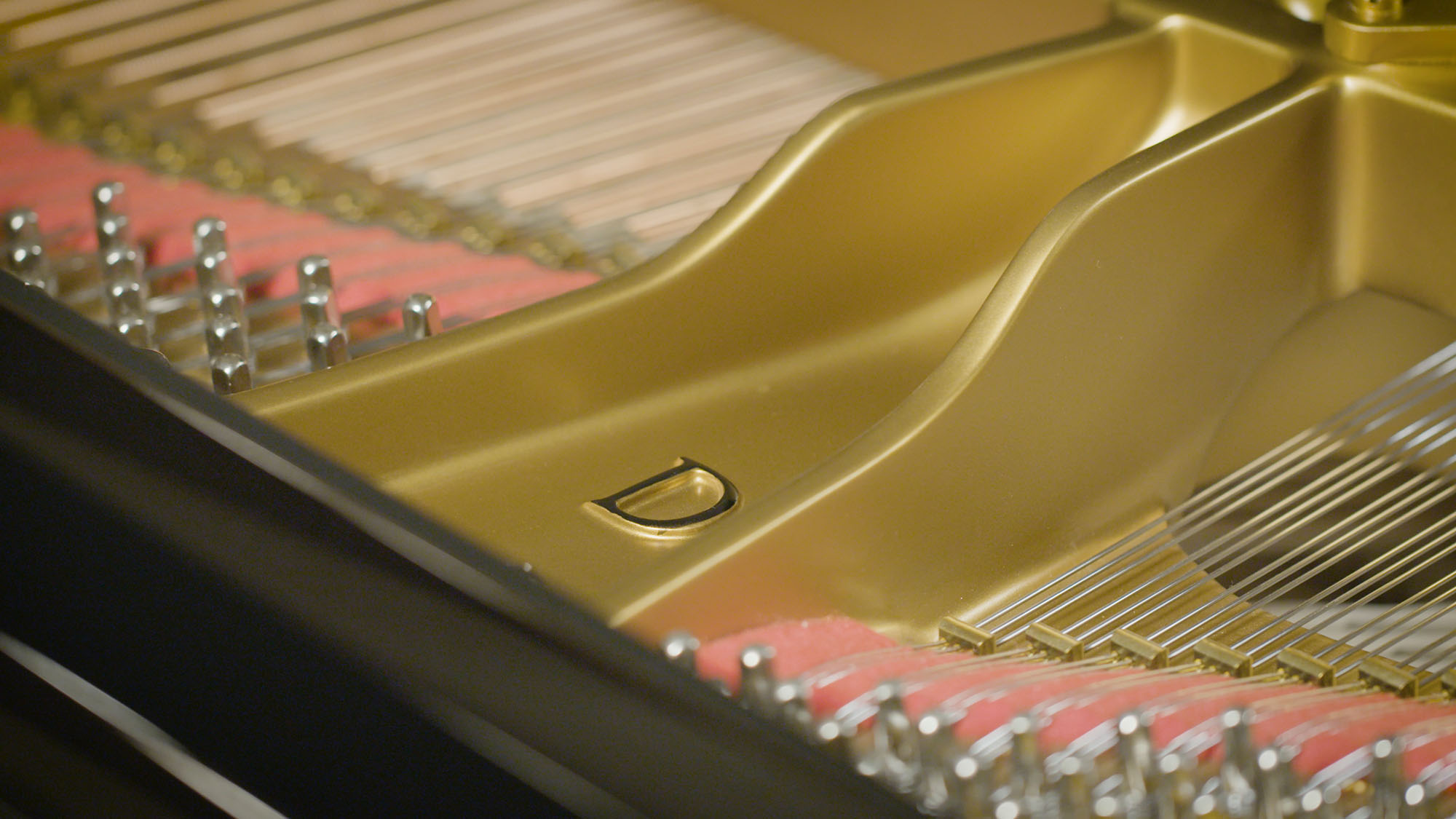 Close-up detailed view of the inside of a grand piano, showing the golden harp frame, steel strings, tuning pins, and red felt dampers. A subtle 'D' logo is visible on the golden frame.