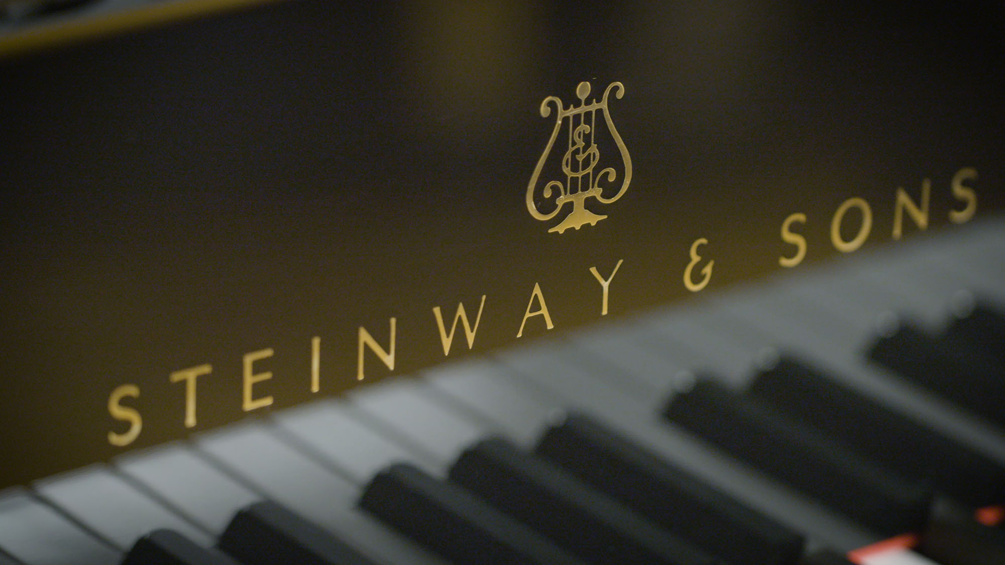 Close-up shot of the Steinway & Sons logo and brand name in gold lettering on a black piano with the white and black piano keys blurred in the foreground.