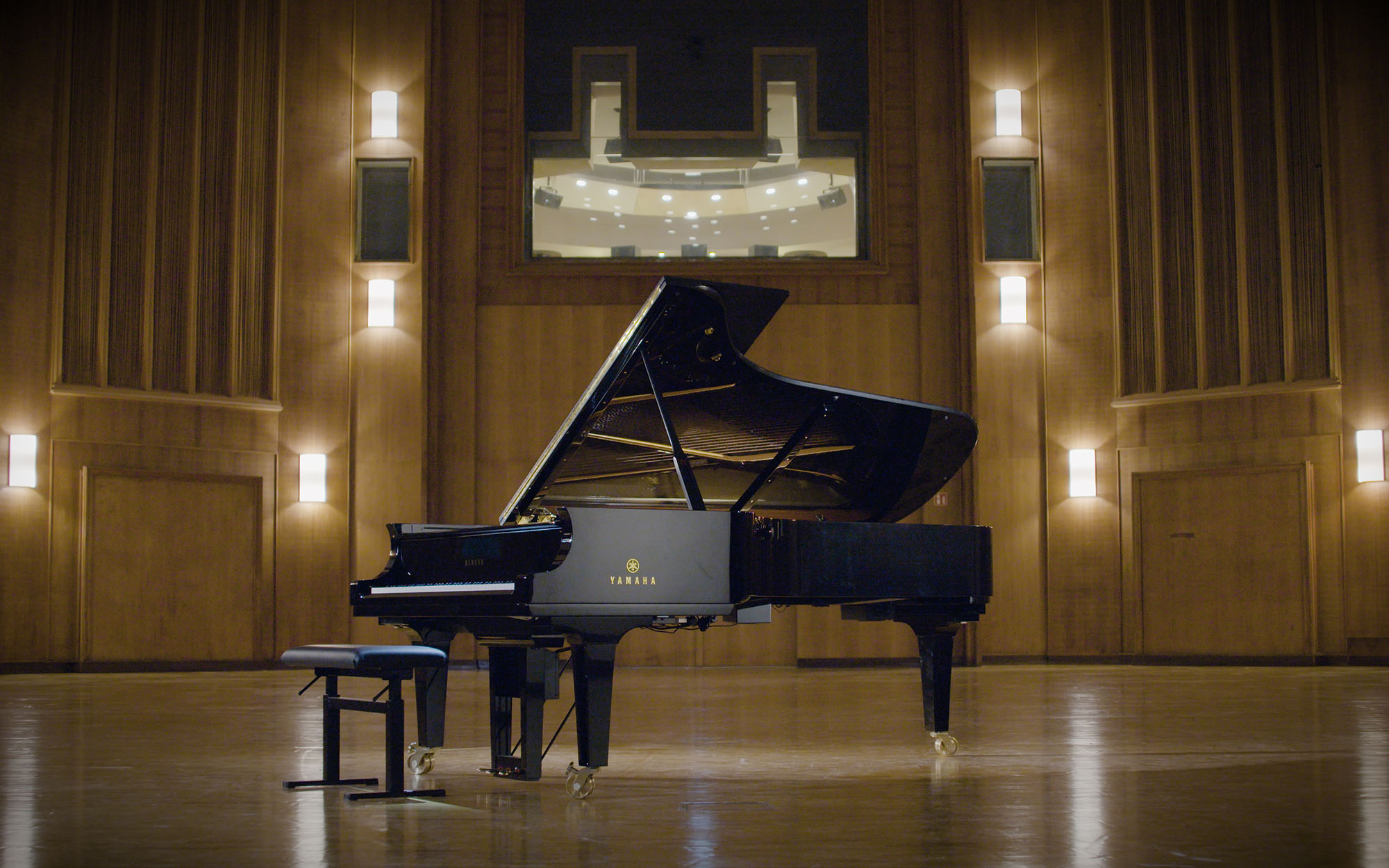 Yamaha grand piano with lid open on a wooden stage in an empty concert hall featuring warm wood paneling, wall sconces, and a large window overlooking a recording studio control room.