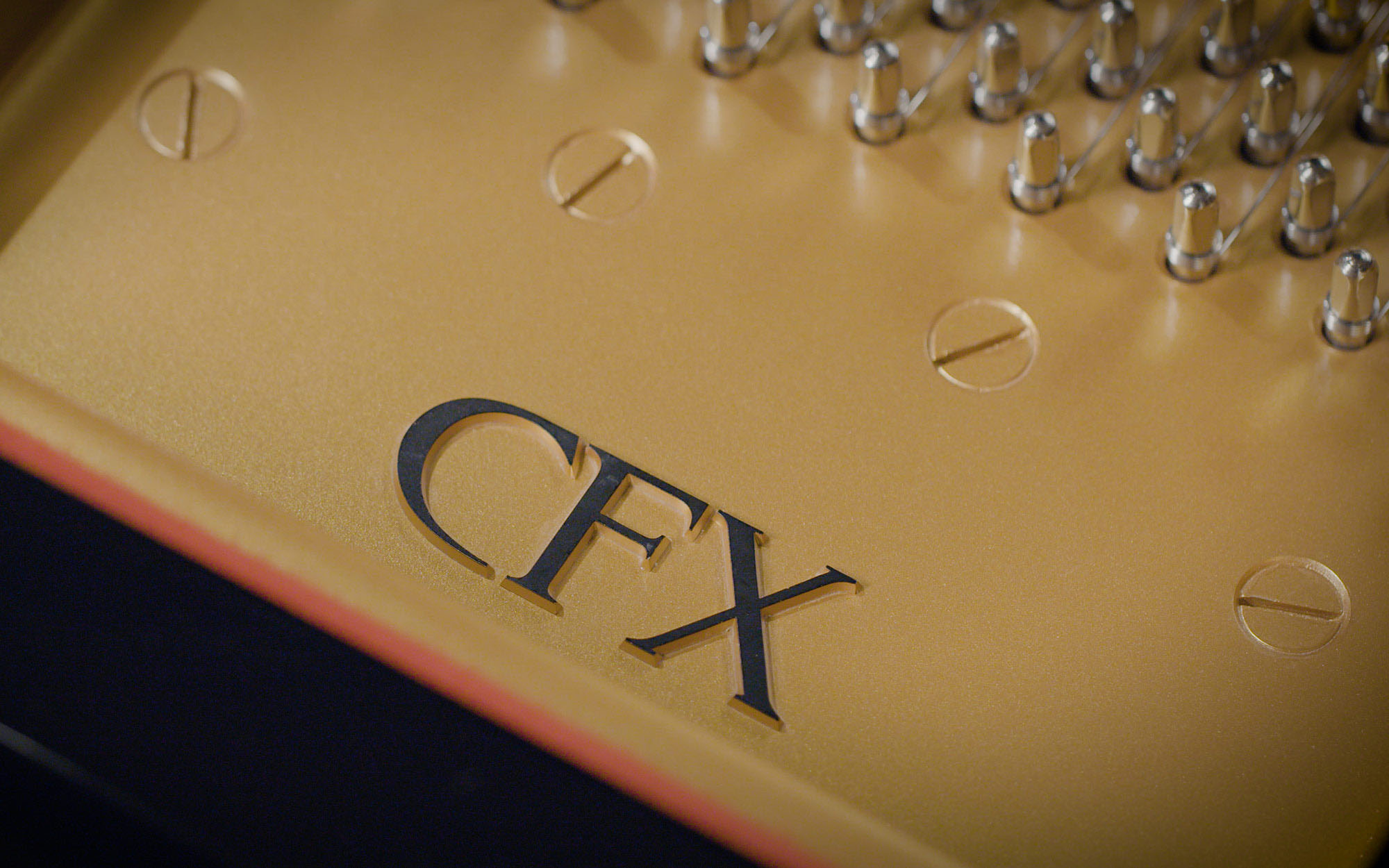 Close-up view of a gold piano soundboard with the black letters CFX
engraved on it, surrounded by silver tuning pins and screws, hinting at the high quality and craftsmanship of a Yamaha CFX concert grand piano.
