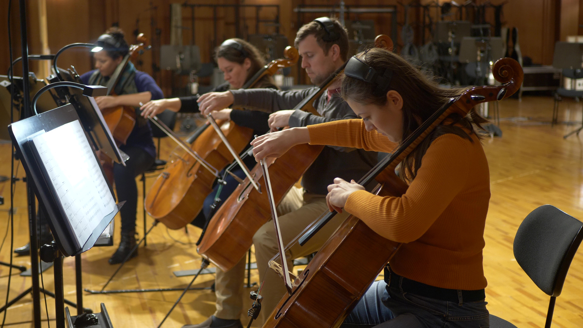 Four musicians, three women and one man, playing cellos in a recording studio, with music stands and sheet music visible.