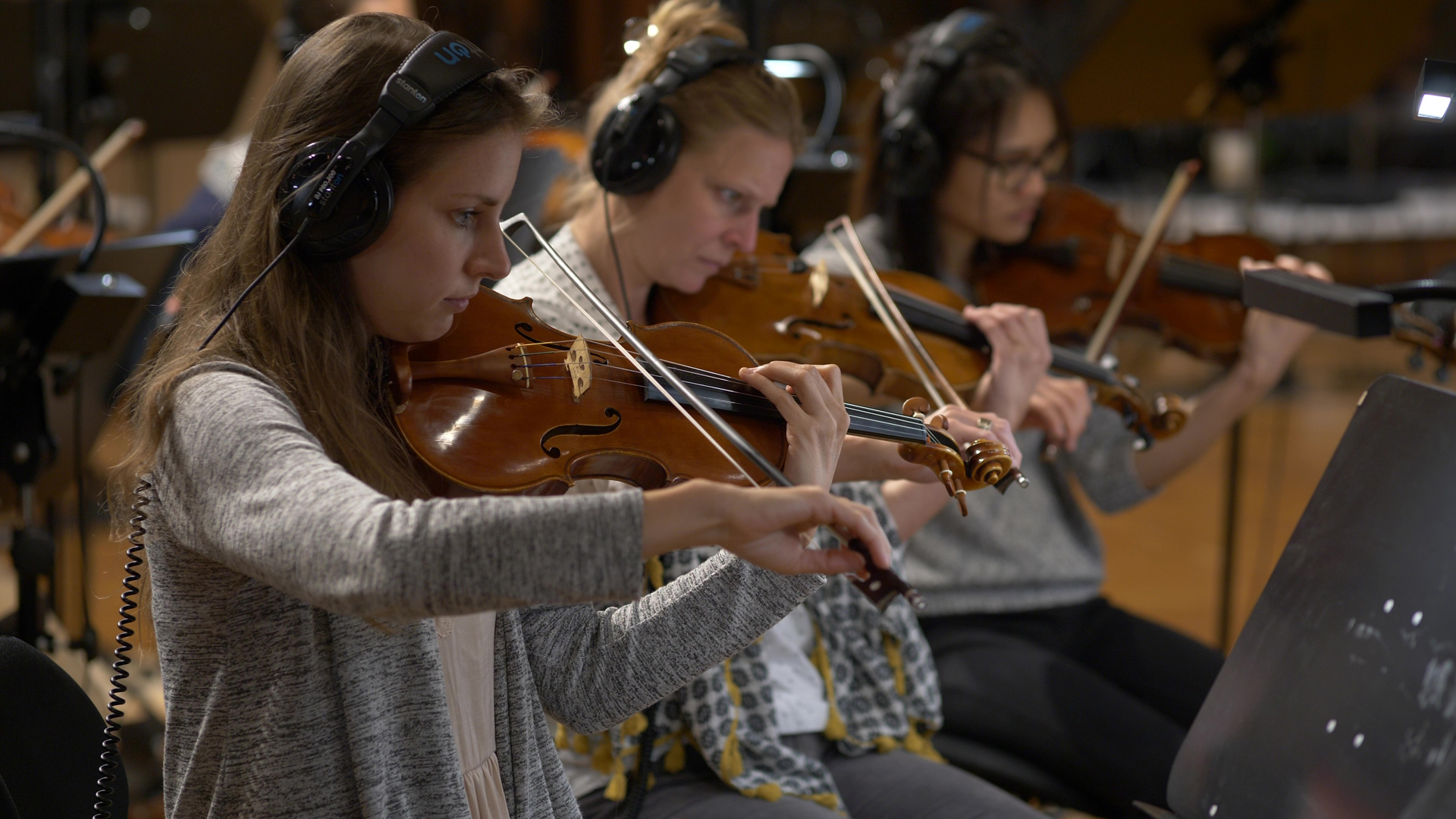 Three female musicians wearing headphones playing violins in a recording studio or orchestra.