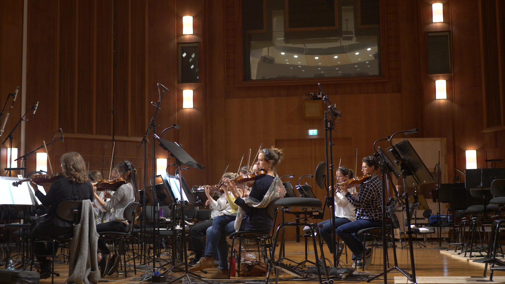 Violin section of an orchestra recording in a studio. Women actively play violins, surrounded by music stands, microphones, and studio recording equipment. The room features warm wooden walls and ambient lighting.